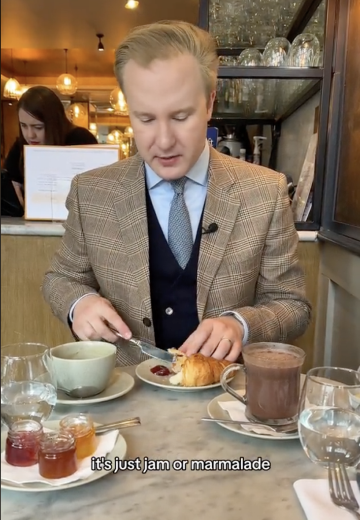 British etiquette expert William Hanson sitting at a table with a croissant, coffee, hot chocolate etc.